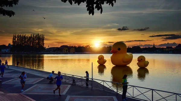 Udon Thani, Tayland - 17 Aralık 2020: İnsanların görüntüleri akşamları gün batımında yayınlanıyor. Arka planda gölde yüzen üç dev sarı lastik ördek, yumuşak odaklı. 