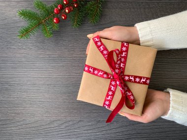 Elinde Noel kurdelesi olan bir hediye kutusu olan bir kız köknar ağacı dallarıyla süslenmiş ahşap bir masada. Düz konum, boşluğu kopyala.