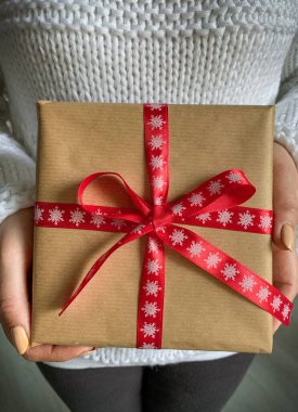 Beyaz kazaklı kadın elinde kırmızı kurdeleli Noel hediyesi kutusu tutuyor..