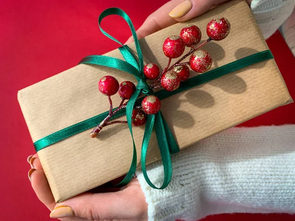Brown craft paper present with green ribbon and branch of berries in hands. Present making for give in holidays or special day.