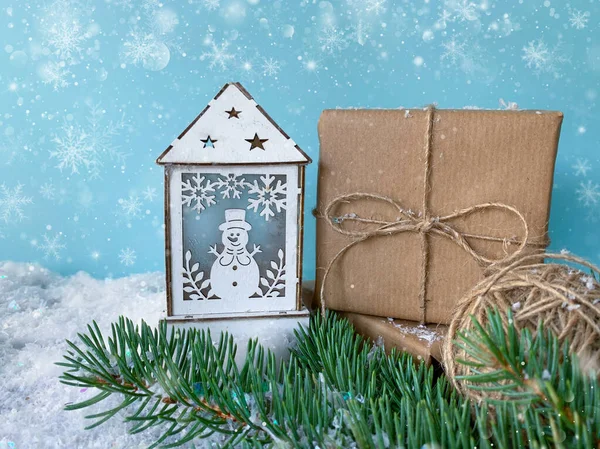 Christmas decorations. Home sweet home. Winter background. White wooden candle lantern with snowman next to handcrafted gift and twine ball over snowy fir tree branch.
