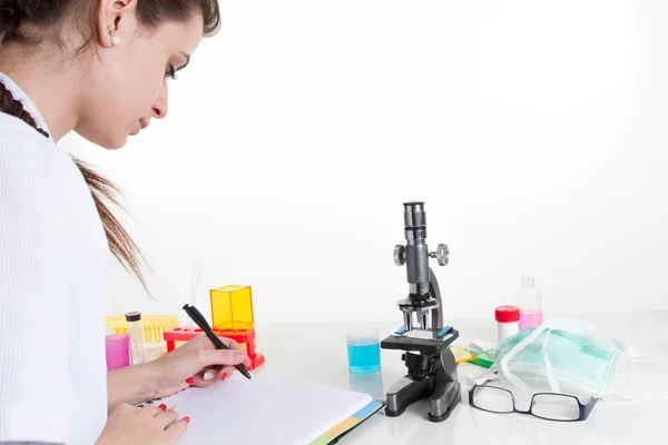 Τον έλεγχο των αποτελεσμάτων. Σοβαρές νεαρός επιστήμονας θηλυκό χρησιμοποιώντας μικροσκόπιο και γραφής στο μπλόκ σημειώσεων ενώ κάθεται στο χώρο εργασίας της — Φωτογραφία Αρχείου