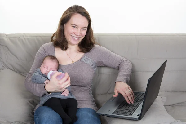 Baby and busy young woman with notebook sit on the sofa in the room.