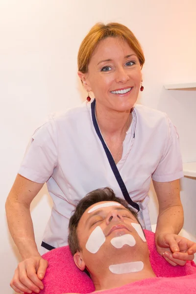 Schöner Mann, der Gesichtscreme von einer blonden Kosmetikerin aufträgt — Stockfoto