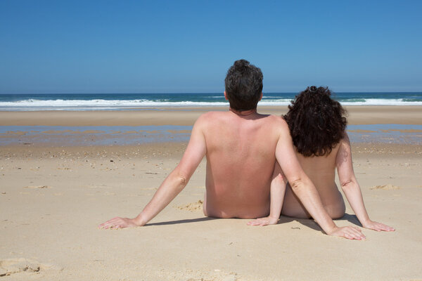 Relaxing nudist couple sitting on beach under deep blue sky