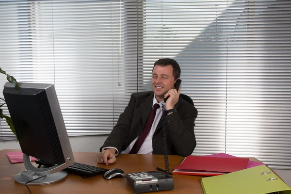 Portrait d'un homme souriant parlant au téléphone, assis au bureau, regardant l'écran de l'ordinateur . — Photo