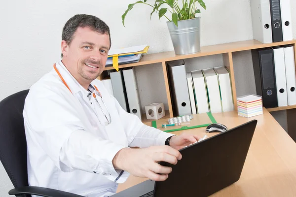 Portrait of a smiling male doctor sitting at desk in medical office — Stock Photo, Image