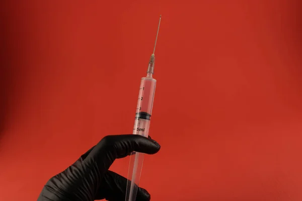 Injection syringe in a black gloved hand close up on a red background