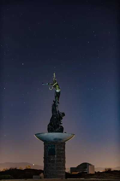 Mermaid statue stands lit against the night sky full of stars and constellations in Autumn night.