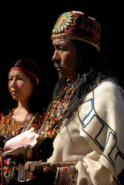 Amerika 'dan iki yerli katılımcı törende ayakta duruyor. Öndeki adam güneş desenli tekstil başlık ve katmanlı tohum-kabuk kolye giyer, arkasında bir kadın ise deniz kabuğu tutar; yan ışık dar bir karşı profillerini oyar