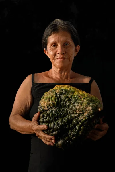 Amerika 'nın yerlilerinden biri büyük, aile yadigarı bir kabağı karanlık bir zemine karşı tutuyor. Ilık yan ışık, ellerindeki meyvelerin dokusunu ve bilgeliği ortaya çıkarır. Atalarının tarım bilgisini onurlandırır ve tohumların devamlılığını sağlar..