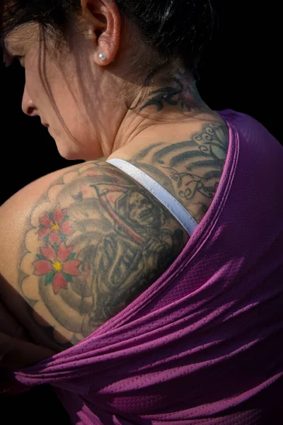 Omzunda ve boynunda Santa Muerte dövmesi olan, mor spor bluzu ve inci küpesi olan bir kadının yakın çekimi. Sert kenar oturumu doku ekler; siyah arkaplan mürekkep ve cilt detaylarını izole eder.