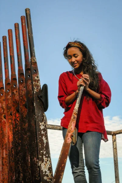 Genç bir kadın, paslı metal ve mavi gökyüzü ile çelişen kırmızı bir bluz ve kot giymiş, eski bir demiryolu kolu yanında duruyor.