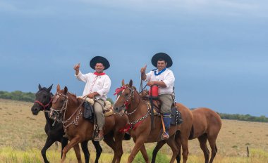 Pampas kırsalındaki geleneksel Arjantin hac yolculuğu sırasında gururla başparmaklarını uzatarak at sırtında yan yana at süren iki beceriksiz..