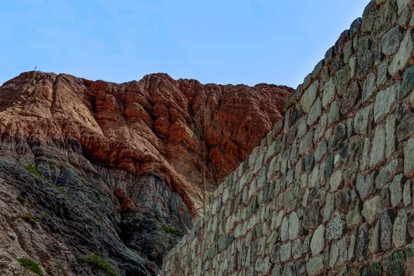 Arjantin Purmamarca 'nın renkli tepelerine doğru uzanan taş bir duvar, temiz bir gökyüzünün altındaki katmanlı mineral dokularını gösteriyor..