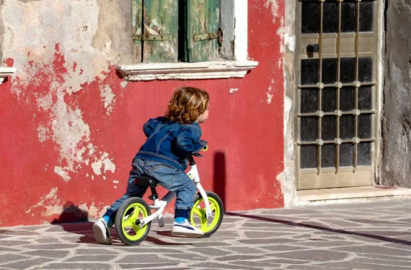 Burano, Venedik, İtalya, Nisan 2019, kıvırcık saçlı komik küçük çocuk dar sokaklarda bisiklet sürüyor.
