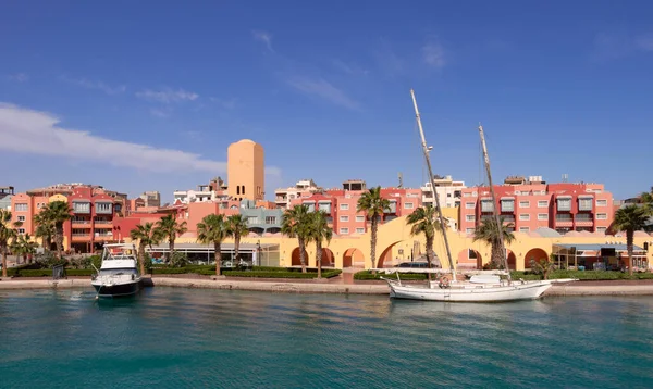 Hurghada marinasından şehrin tepesine kadar panoramik bir manzara. Dükkanlar, kafeler ve yürümek için yayalar, ön planda demirli bir yelkenli yat ve bir motorlu tekne var.
