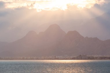 Günbatımında ünlü Türk Konyaalti Sahili manzarası. Güneş ışınları bulutları delip geçer ve dağları aydınlatır. Antalya, Türkiye