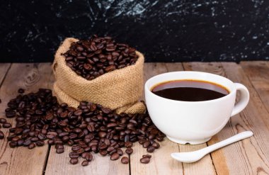 Koyu kahve ve bir kaşık ile beyaz kahve fincanı eski ahşap zeminde duran bir torbada kokulu kahve çekirdekleri vardı..