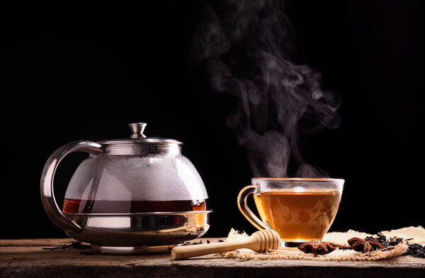 A cup of tea and hot steam from brewed tea with a glass jug with herbs placed on a black background wooden table.