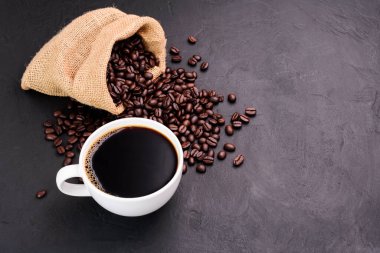 White coffee cup and coffee beans in a sack on dark background, top view.