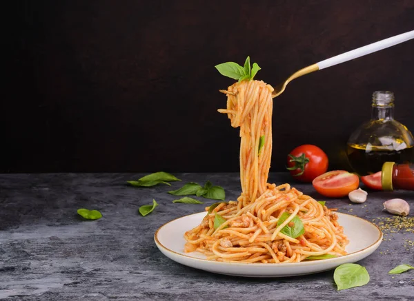 Çatal, ağız sulandıran domates ve fesleğen soslu klasik İtalyan spagetti makarnasını topluyor. Taze pişmiş yiyecekler ahşap bir masada bir tabak sete konur. koyu bir arkaplanda