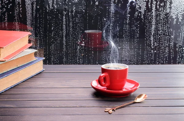 Yağmurlu bir günde cam zeminde yağmur damlaları ve pencerenin kenarındaki kitaplarla dolu eski bir ahşap masaya kurulmuş sıcak siyah kahve fincanı..