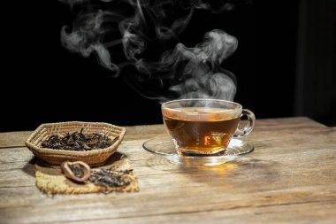 Bardakta sıcak çay dumanlı ve ahşap bir masada kuru çay yapraklı..