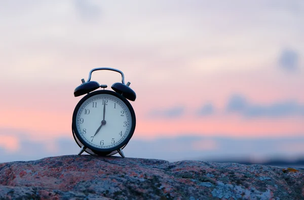 Un reloj despertador negro en roca a las siete en punto al aire libre durante el amanecer . —  Fotos de Stock