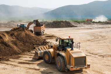 Dağlık bir bölgede, bir yaz gününde toprak işleri. Tekerlek yükleyiciler, buldozerler ve çöp kamyonları çalışıyor. Bu toprağı taşıyan çöp kamyonlarının tozu.