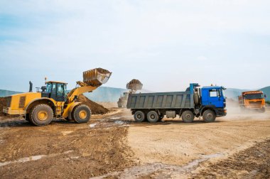 Dağlık bir bölgede, bir yaz gününde toprak işleri. Tekerlek yükleyiciler, buldozerler ve çöp kamyonları çalışıyor. Bu toprağı taşıyan çöp kamyonlarının tozu.