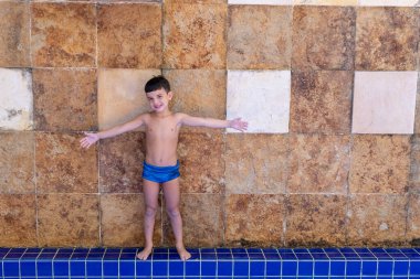 6 yaşında çocuk havuzun kenarında kollarını açmış, arka planda taş bir duvar var (fotoğraf 1).