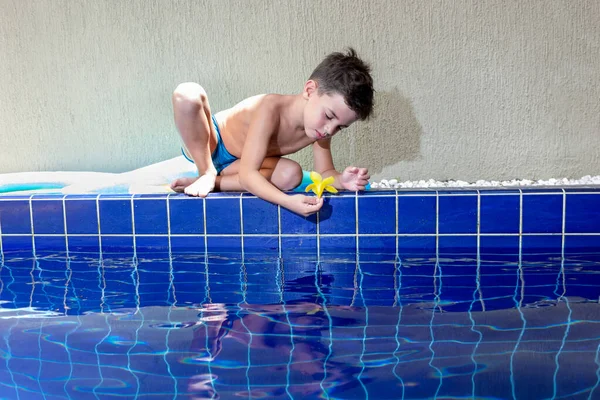 6 yaşında bir çocuk havuzun kenarında oturmuş sarı bir çiçekle oynuyor..