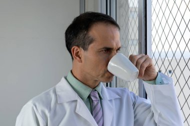 44 yaşında beyaz önlüklü, kravatlı, kahve içen ve pencereden dışarı bakan bir doktor..