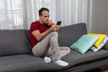 Olgun bir adam (44 yaşında) kanepede çoraplarıyla oturuyor ve akıllı telefonuyla oynuyor..