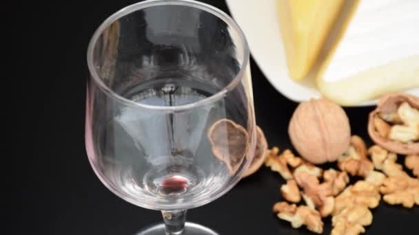 Primer plano de una copa de vino tinto que se vierte. En el fondo desenfocado, frutos secos y parmesano y brie (zoom en). — Vídeos de Stock
