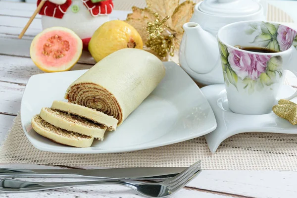 Bolo de rolo (rulo kek) yanında çatal bıçak, kahve ve guava dilimleri.
