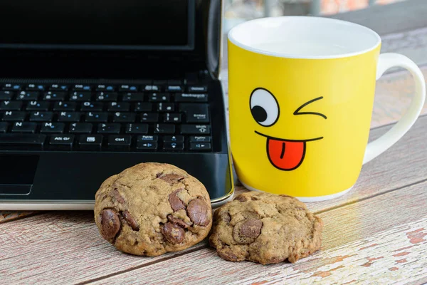 Cookies with chocolate drops leaning on the laptop next to a yellow mug with milk, break for e-learning (side view).