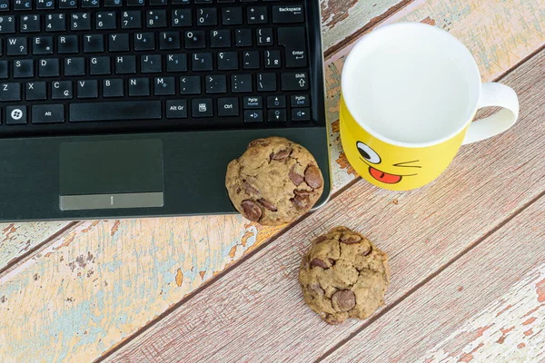 Cookies with drops of chocolate on the laptop, next to a yellow mug with milk, break for e-learning (top view).