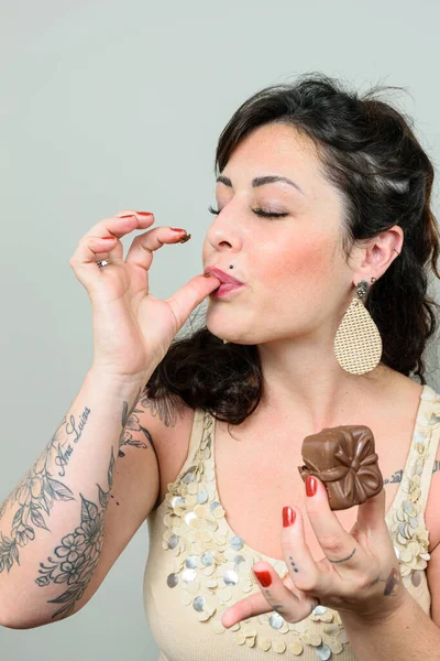 Tattooed woman, with her eyes closed, licking her fingers and holding a Brazilian honey cake.