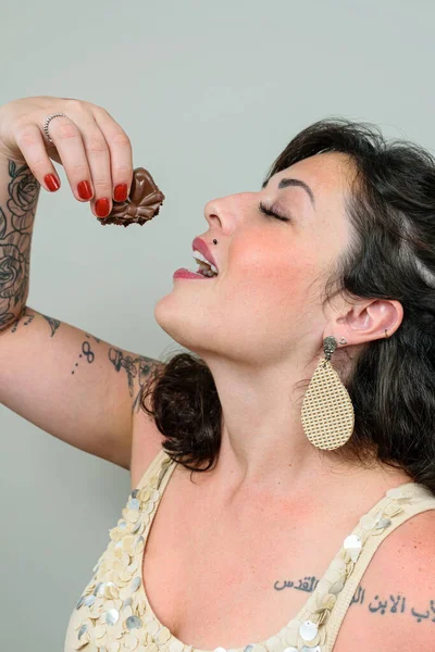 Tattooed woman with mouth open and eyes closed, holding a Brazilian honey cake above her head. Tattoo on left shoulder written in Arabic - Father, Son and Holy Spirit.