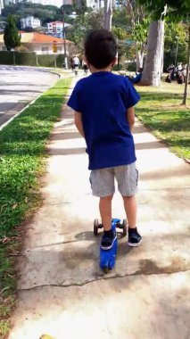 Güneşli bir günde, halka açık bir meydanda scooterıyla yürüyen 8 yaşında bir çocuk.).