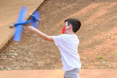 8 yaşındaki çocuk, maske takıyor, halka açık bir meydanda yapay köpük uçağıyla oynuyor (yan görünüm)).