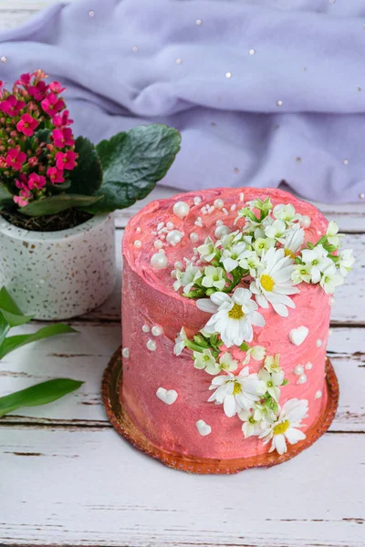 Pembe tereyağlı kremayla kaplı çikolatalı pasta. Beyaz kalpler, inciler ve çiçeklerle süslenmiş. Arka planda mor bir kumaş.