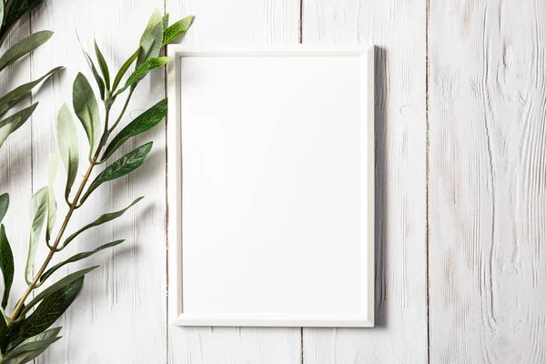 White photo frame with a sprig of eucalyptus on a white wooden table. Minimalistic, gentle mock up