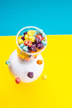 Mavi-sarı arka planda şeffaf bir bardakta renkli patlamış mısırlı yaratıcı kompozisyon. Kopyalama alanı olan modern soyut yaşam