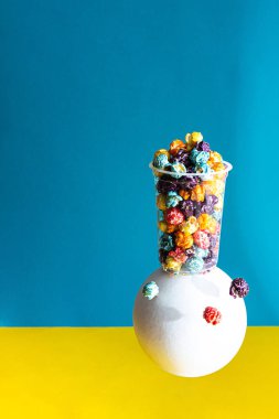 Mavi arka planda şeffaf bir bardakta renkli patlamış mısırlı yaratıcı kompozisyon. Beyaz küreli modern natürmort yaşam ve kopyalama alanı