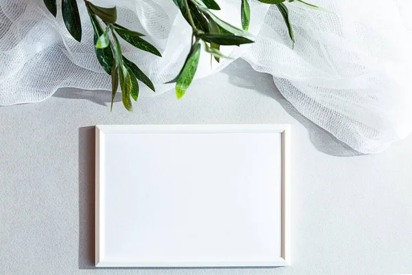 White blank photo frame on a gray-white background, with an olive branch. Modern minimal still life, top view. Mock up
