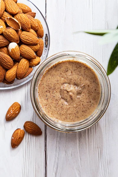 Bir kavanoz badem ezmesi ve beyaz ahşap bir masada zeytin dalı ile bir demet taze badem. Ev yapımı nişasta yağı, doğal ve sağlıklı yiyecekler. Modern sağlık ve vejetaryen konsepti. 