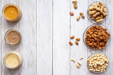 Modern refah ve veganlık kavramı. Beyaz ahşap bir masada kavanozlar dolusu badem, fıstık ezmesi ve fıstık ezmesi. Sağlıklı bir kahvaltı için organik, ev yapımı fıstık ezmesi..